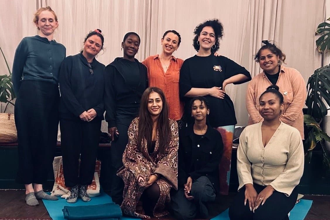A group of nine women smiling together after a workshop, some standing and some seated on yoga mats